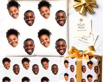 Könnte beinhalten: Weißes Geschenkpapier und eine Geschenkbox mit sich wiederholenden Bildern von lächelnden Gesichtern. Eine goldene Schleife und ein Geschenkanhänger mit handschriftlichem Text sind ebenfalls vorhanden. Die Geschenkbox ist mit einem goldenen Band gebunden.