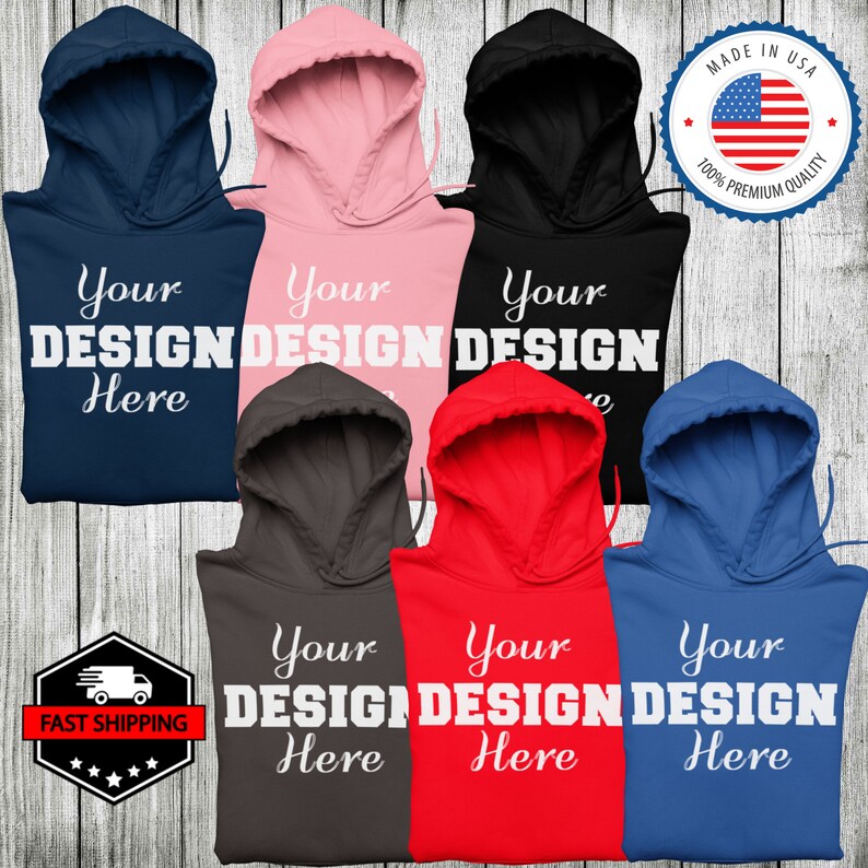 May include: Six different color hooded sweatshirts with the text "Your Design Here" printed in white on the front. The sweatshirts are arranged in a semi-circle on a woodgrain background.