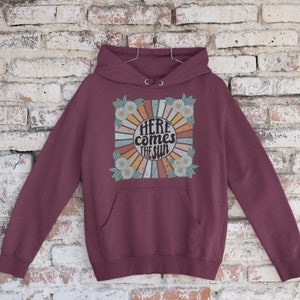 Op de afbeelding: Bordeauxkleurige hoodie met een retro-grafische print van een zon met de tekst "Here comes the sun" omringd door madeliefjes.