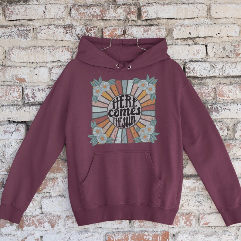 Op de afbeelding: Bordeauxkleurige hoodie met een retro-grafische print van een zon met de tekst "Here comes the sun" omringd door madeliefjes.
