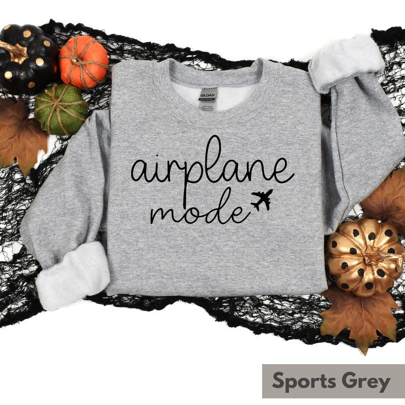 Op de afbeelding: Een grijs sweatshirt met de tekst "airplane mode" in zwarte cursieve letters en een klein vliegtuigpictogram. Het sweatshirt ligt op een zwarte kanten achtergrond met herfstdecoraties.