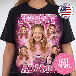 May include: Black t-shirt featuring a graphic of a young woman with pink headphones, a pink hoodie, and a gaming controller. The text "JENNY ADAMS" is displayed in pink, with a "Made in USA" graphic.