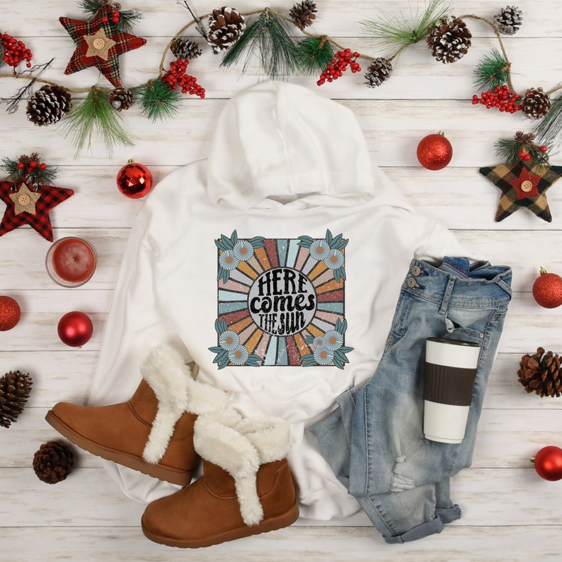 Op de afbeelding: Witte hoodie met een retro-grafische print met een zonnestralenontwerp en de tekst "Here comes the sun". De grafiek is omgeven door een bloemige rand.