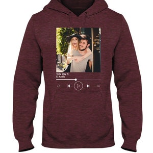 May include: A maroon hoodie with a photo of a couple on the front. The photo is framed with a white border and has a music player interface below it. The music player interface shows the song title "Te la Doy" by "El Ankla".