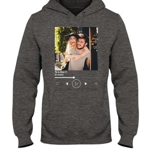 May include: A gray hoodie with a photo of a couple on the front. The photo is in a white frame with a music player interface below it. The music player interface shows the song title "Te la Doy" by "El Ankla" and play, rewind, and fast forward buttons.