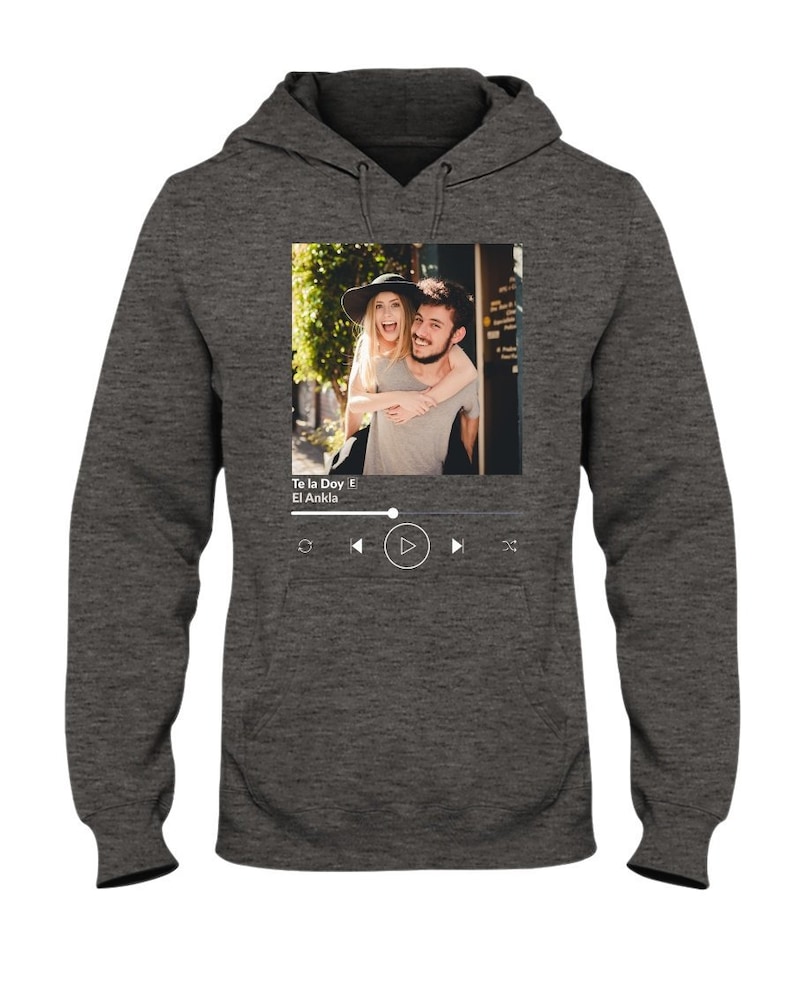 May include: A gray hoodie with a photo of a couple on the front. The photo is in a white frame with a music player interface below it. The music player interface shows the song title "Te la Doy" by "El Ankla" and play, rewind, and fast forward buttons.