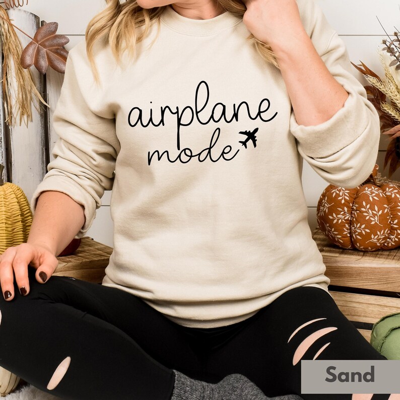 Op de afbeelding: Een beige sweatshirt met de tekst "airplane mode" en een kleine vliegtuigafbeelding gedrukt op de voorkant. Het model draagt ​​zwarte leggings.