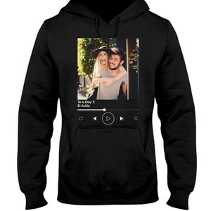 May include: Black hoodie with a graphic of a couple in front of a building. The graphic includes a music player with the song title "Te la Doy" by "El Ankla" displayed.