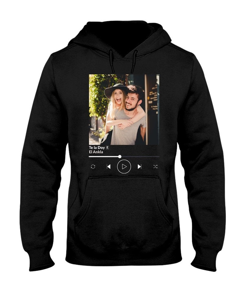 May include: Black hoodie with a graphic of a couple in front of a building. The graphic includes a music player with the song title "Te la Doy" by "El Ankla" displayed.