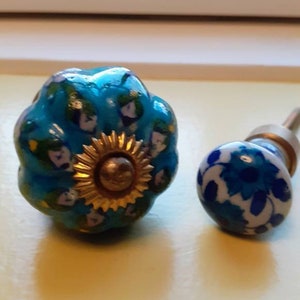 Puede incluir: Dos tiradores decorativos para armarios. Uno es azul turquesa con un diseño de girasol dorado. El otro es blanco con motivos florales azules. Estos tiradores son probablemente para muebles o decoración del hogar.