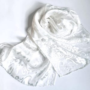 May include: A white scarf featuring a tonal, semi-sheer damask pattern with swirling floral and leaf motifs. The fabric has a subtle sheen and a lightweight, flowing texture, arranged in a loose, draped coil on a plain white surface.