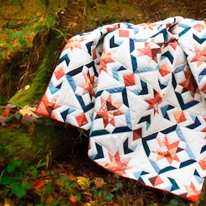Könnte beinhalten: Eine weiße, blaue und orangefarbene Patchwork-Decke mit Sternenmuster liegt auf einem moosbewachsenen Baumstamm in einem Wald. Die Decke ist aus verschiedenen Stoffen gefertigt und hat einen rustikalen, handgefertigten Charakter.