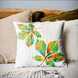 Könnte beinhalten: Ein weißes quadratisches Kissen mit einem farbenfrohen Blattdesign. Die Blätter bestehen aus geometrischen Formen in Grün, Orange und Gelb. Das Kissen liegt auf einem Bett mit anderen Kissen und einer grauen Decke.
