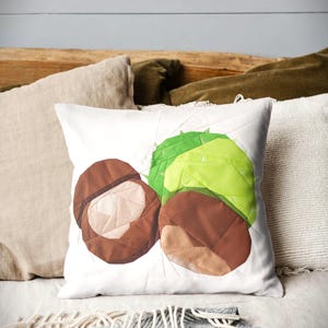 Könnte beinhalten: Weißes quadratisches Kissen mit einem Patchwork-Design aus braunen Kastanien und einer grünen, stacheligen Hülle. Das Kissen liegt auf einem Bett mit anderen Kissen und einer grauen Decke mit geometrischem Muster.