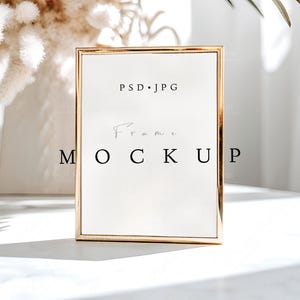 May include: A gold-framed rectangular frame mockup with the text "PSD.JPG" at the top and "Frame Mockup" in the center. The frame is on a white surface, with soft lighting and decorative elements in the background.