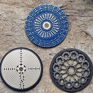Könnte beinhalten: Drei runde dekorative Untersetzer mit komplizierten Designs. Der erste Untersetzer hat ein blaues und weißes geometrisches Muster. Der zweite Untersetzer hat ein weißes Labyrinth-Design. Der dritte Untersetzer hat ein schwarzes und silbernes Blumenmuster.