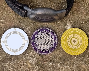 May include: Three round, decorative pendants with intricate designs. One is white with gold accents, another is purple with a geometric pattern, and the third is yellow with a radial design. A black strap with a gray clasp is visible at the top.