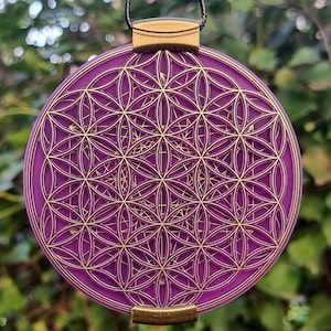 Può includere: Un ciondolo rotondo di colore viola con un design dorato a forma di fiore della vita. Il ciondolo è rotondo e ha un'anellina dorata.