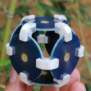 May include: A blue and gold geometric sphere made of interlocking circular pieces. The sphere is held in a hand and has a white plastic frame.