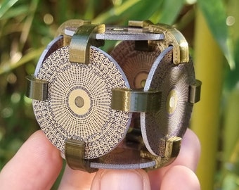 Può includere: Un cubo di metallo dorato con un motivo geometrico su ogni lato. Il cubo è composto da sei pezzi circolari che sono collegati da barre di metallo.