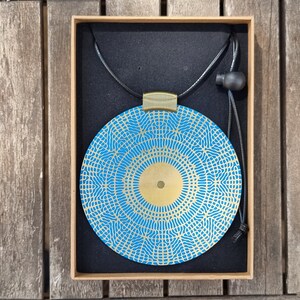 May include: A blue and gold circular pendant necklace with a geometric pattern. The pendant is in a box with a black velvet lining.
