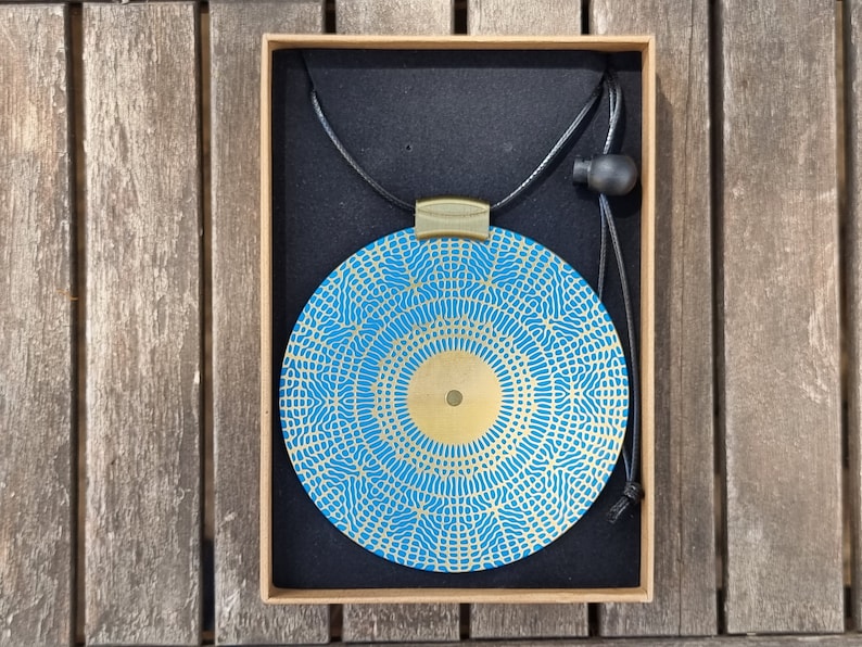 May include: A blue and gold circular pendant necklace with a geometric pattern. The pendant is in a box with a black velvet lining.