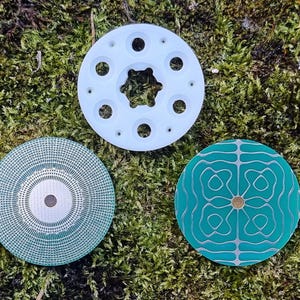 May include: Three circular objects with intricate designs rest on a bed of green moss. One is white with circular holes, another is teal with a geometric pattern, and the third is teal with a dotted pattern.