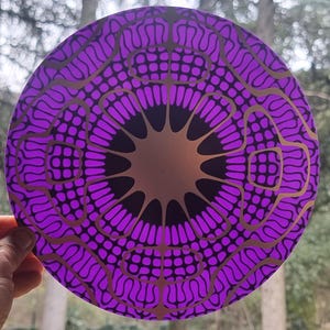Peut inclure: Un mandala rond, violet et or avec un centre noir. Le mandala est composé de motifs et de dessins complexes.