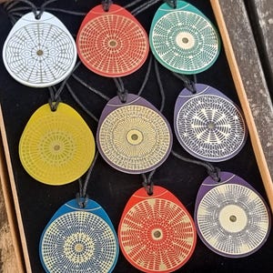 Può includere: Un set di nove ciondoli colorati con intricati disegni geometrici. Ogni ciondolo ha la forma di una lacrima e presenta un motivo diverso in tonalità di bianco, nero, rosso, giallo, verde e blu.