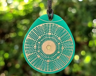 May include: A teal pendant with a gold-colored, intricate circular design. The pendant is suspended from a black cord. The background is a blurred view of green foliage. The pendant has a unique, artistic appearance.