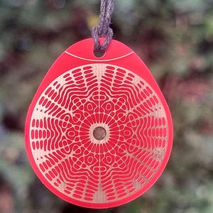 Peut inclure: Un pendentif rouge avec un motif de mandala argenté. Le pendentif est suspendu à un cordon brun.