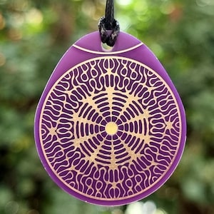 Puede incluir: Un colgante morado con un intrincado diseño circular dorado. El colgante está suspendido de un cordón negro. El fondo es una vista borrosa de follaje verde.