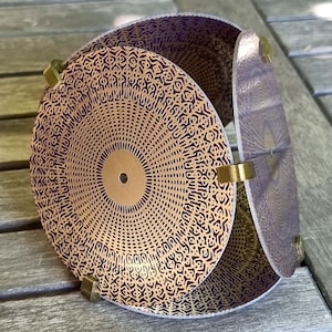 Puede incluir: Una esfera decorativa hecha de múltiples piezas circulares de papel con un patrón geométrico en tonos dorados y morados. La esfera se mantiene unida con bisagras de latón.