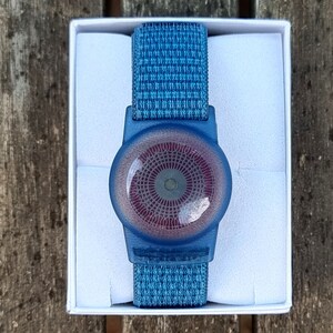 May include: A blue and red circular device with a textured pattern, attached to a blue woven wristband. The device is in a white box.