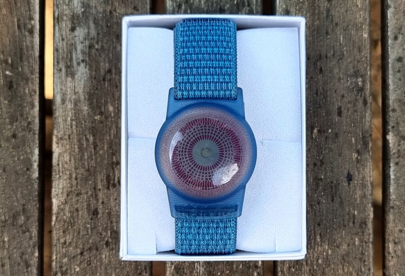 May include: A blue and red circular device with a textured pattern, attached to a blue woven wristband. The device is in a white box.