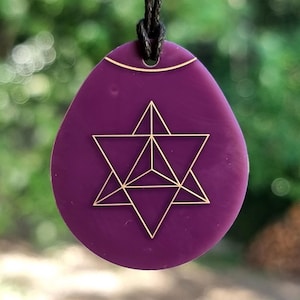 May include: A purple pendant with a gold geometric design, hanging from a black cord. The pendant is oval-shaped with a gold accent at the top. The background is a blurred view of green foliage.