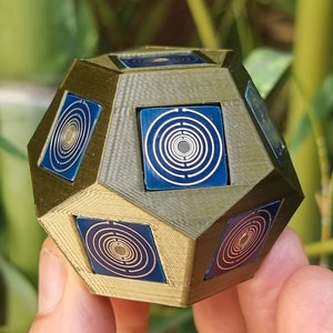Pode incluir: Uma forma geométrica impressa em 3D de cor dourada com faces com padrões azuis e dourados. A forma é um dodecaedro, uma figura de 12 lados.