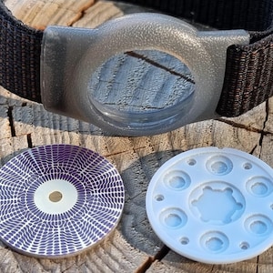 May include: A black nylon strap with a gray circular plastic housing, accompanied by a circular insert featuring a purple and white radial wave pattern and a light gray circular component with multiple recessed circular indentations.