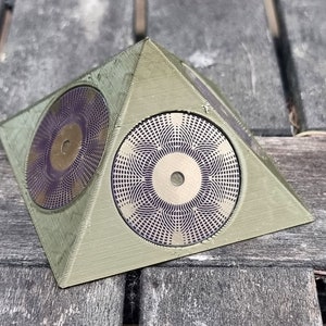 Könnte beinhalten: Ein grünes pyramidenförmiges Objekt mit drei kreisförmigen Designs auf jeder Seite. Die Designs sind eine Kombination aus Gold und Lila.