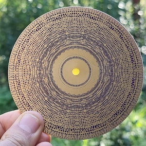May include: A circular, gold-colored object with a detailed, patterned design in dark brown. The pattern consists of concentric circles and intricate lines. A small, yellow circle is in the center. The object is held against a blurred green background.