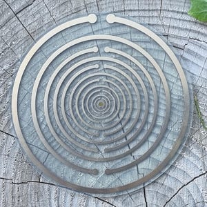 Può includere: Un oggetto metallico argentato, circolare, con anelli concentrici a forma di spirale o bersaglio, poggia su una superficie di legno grigio e invecchiato. Foglie verdi sullo sfondo, creando un contrasto naturale con il design metallico.