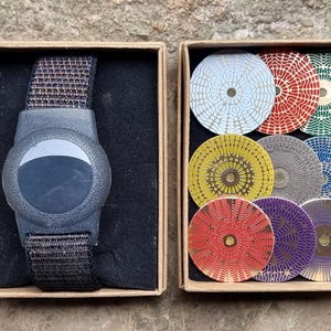 Op de afbeelding: Twee kartonnen dozen op een getextureerd oppervlak. Eén doos bevat een donkergrijs horloge met een zwarte band. De andere doos bevat tien kleurrijke, cirkelvormige objecten met ingewikkelde patronen in verschillende kleuren, waaronder blauw, rood en groen.