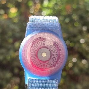 May include: A blue and red circular device with a patterned design, attached to a blue and white strap. The device has a small white circle in the centre.