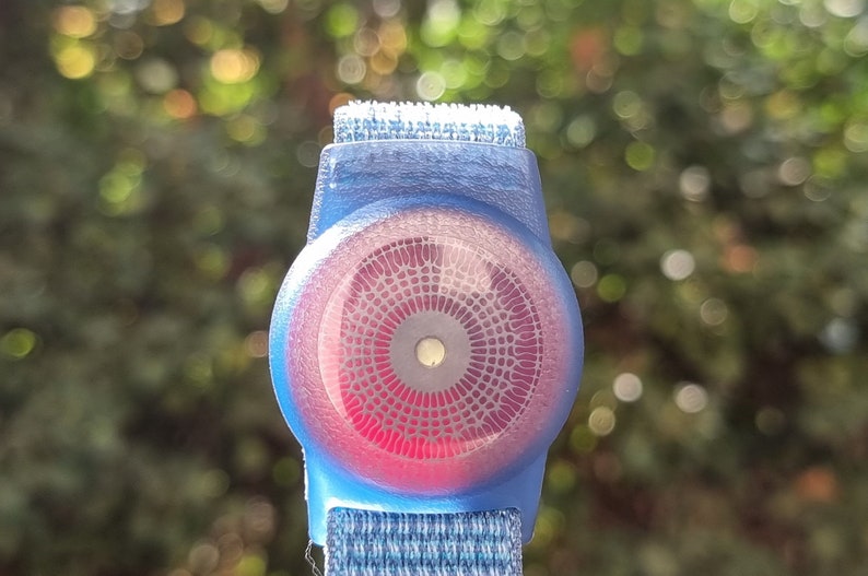 May include: A blue and red circular device with a patterned design, attached to a blue and white strap. The device has a small white circle in the centre.