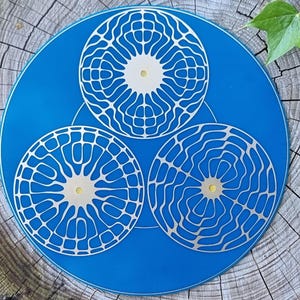 Puede incluir: Una pieza decorativa azul con tres diseños circulares blancos con patrones intrincados. Los círculos están dispuestos en una formación triangular. La pieza está sobre un fondo de madera natural.