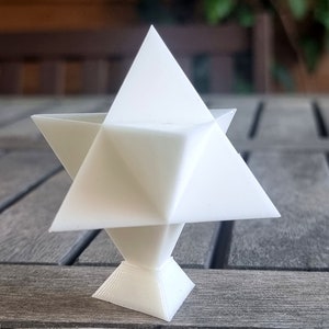 Könnte beinhalten: Eine weiße, dreidimensional gedruckte geometrische Sternform mit einer Basis. Der Stern besteht aus mehreren Dreiecken, die sich zu einer komplexen Form schneiden.