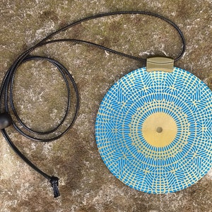 May include: A round blue and gold pendant necklace with a geometric pattern. The pendant is attached to a black cord.
