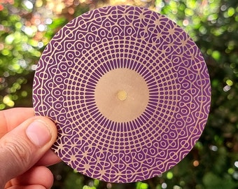 May include: A round, purple and gold mandala design on a coaster. The coaster has a geometric pattern with a central circle.