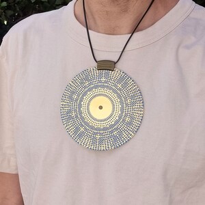 May include: A gold pendant necklace with a circular design featuring a blue and yellow geometric pattern. The pendant has a small gold circle in the center.