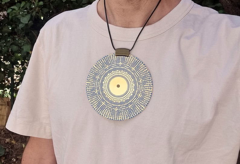 May include: A gold pendant necklace with a circular design featuring a blue and yellow geometric pattern. The pendant has a small gold circle in the center.
